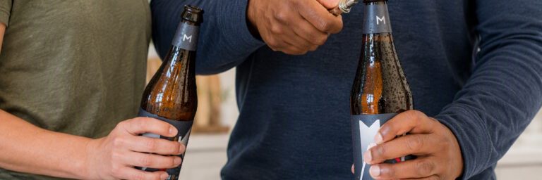Two people in casual shirts clinking brown beer bottles with condensation on the glasses.